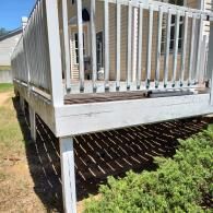 A white wooden deck with vertical railings stands elevated above a yard with grass and a low-lying green shrub.