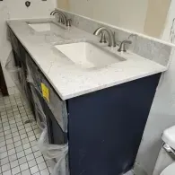 A double-sink bathroom vanity with a white marble countertop and dark blue cabinetry, partially wrapped in plastic.