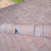 A close-up view of a shingled roof showing a small hole or tear in one of the ridge cap shingles.