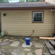 A beige, wood-sided house exterior with a single window and a blue bucket resting on a stone patio.