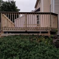 A newly constructed wooden deck with railings attached to the back of a light-colored house, overlooking a green shrub.