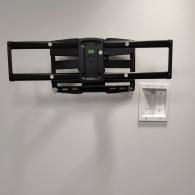 A black articulating TV wall mount is installed on a white wall next to a recessed white cable management outlet.