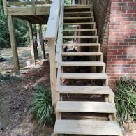 A newly constructed wooden outdoor staircase with a handrail leading up to a deck beside a brick wall.