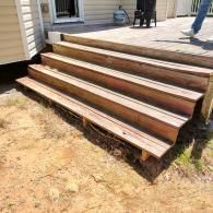 Wooden steps lead up to a deck attached to a house, with sandy ground in the foreground and a person’s legs visible above.