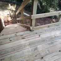 A high-angle view of a wooden staircase leading down from a deck to a dirt-covered yard, featuring wooden railings.