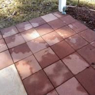 Red square patio pavers with damp patches from rain, adjacent to a white post and grassy area.