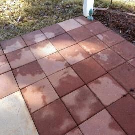 A patio made of square, red concrete pavers partially wet with rain or melting snow, positioned next to a grassy area.