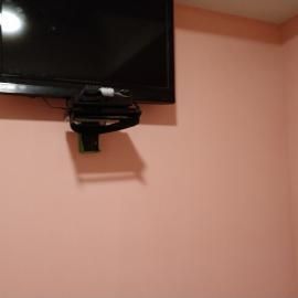 A television mounted on a pink wall with a black device placed on a shelf attached directly underneath the screen.