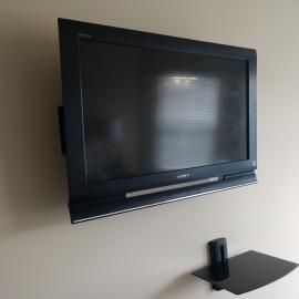 A black flat-screen television mounted on a plain, light-colored wall with a small, black floating shelf below it.