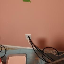 A white wall outlet with multiple black and white power cords plugged into it, above a paint tray and drop cloth.
