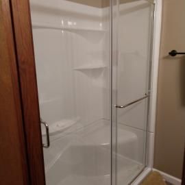 A white shower stall with a built-in corner seat and glass sliding doors in a bathroom with beige walls.