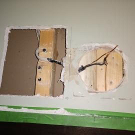 Two holes cut in a drywall ceiling show wooden studs and electrical wiring.