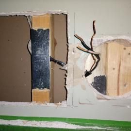 A hole in a drywall section showing two internal wall cavities, wooden studs, a metal guard, and exposed electrical wires.