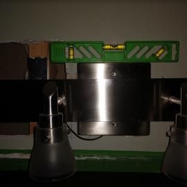 A level tool rests horizontally on top of a metallic wall-mounted light fixture in a dim room.