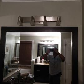 A person holds a phone to take a mirror selfie in a bathroom with a four-light fixture and dark wood-framed mirror.