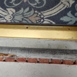 A view looking down at a doorway threshold, showing a patterned carpet, a gold-colored metal strip, and a damaged floor.