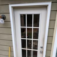 A white-framed door with twelve glass panes, set in a tan-sided wall next to an outdoor light fixture.