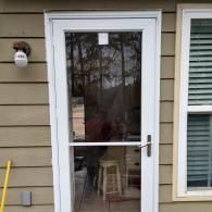 A white storm door with a glass panel on the side of a house with tan horizontal siding and an outdoor light fixture.