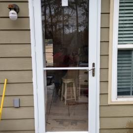 A white storm door with a central metal handle is set against beige horizontal siding, next to a window and a wall light.