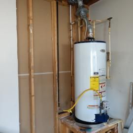 A gas water heater standing in an unfinished room with exposed wooden studs and plumbing.