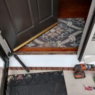 A partially open door with a metal sweep being installed, resting on a welcome mat with a drill nearby on the porch.