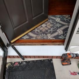 A brown door slightly open over a floral rug, with a door closer, drill, and welcome mat on the exterior porch.