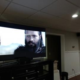 A television screen displaying a man with a beard, set in a room with a drop ceiling and wall molding.
