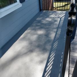 A light grey porch floor next to a blue wall and black metal railing, with tree shadows cast across the surface.