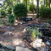 A backyard garden landscape featuring a small pond, rock borders, and a black water filtration unit near dense trees.