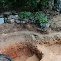 An excavated dirt pit with layers of reddish soil and a rocky, landscaped border with green plants above.