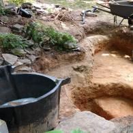 A black pre-formed pond liner sits beside a partially excavated dirt pit in a rocky outdoor setting with a wheelbarrow.