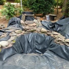 An outdoor pond under construction, lined with a black tarp and surrounded by scattered grey rocks in a garden setting.