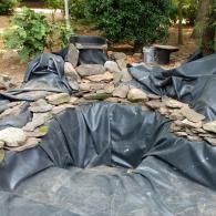An unfinished garden pond lined with black rubber sheeting, surrounded by scattered rocks in a backyard setting.
