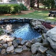 A stone-lined garden pond with dark, rippling water sits in a backyard near a grassy lawn and a deck.