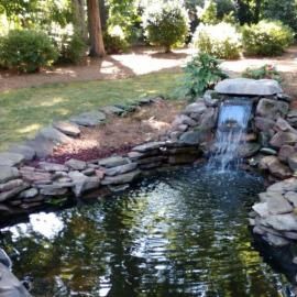 A stone-lined garden pond featuring a small waterfall, surrounded by trees and lush greenery on a sunny day.