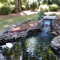 A backyard pond with a small rock waterfall, surrounded by stones and lush green landscaping.