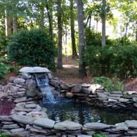 A small stone waterfall flowing into a landscaped garden pond surrounded by tall trees and lush greenery.