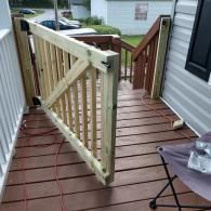 A wooden safety gate stands open on a brown deck next to stairs, a house wall, and a folding chair.