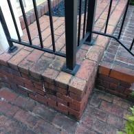 A brick porch corner featuring a black metal railing base mounted on a weathered, reddish-brown brick edge.