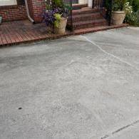 A concrete driveway with a thin, irregular crack leading toward a brick house entrance with stairs and potted plants.