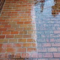 A brick surface being power washed, showing a clean, bright orange side on the left and a dirty, dark side on the right.