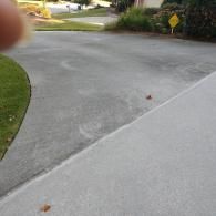 A concrete driveway with circular patterns from pressure washing, beside a patch of green grass and a small yellow sign.