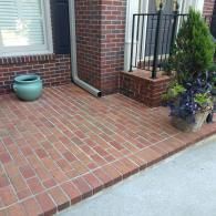 A brick porch with a green ceramic pot on the left and a potted evergreen tree by the brick stairs on the right.