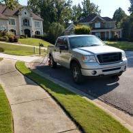A silver pickup truck parked on a residential street with a hose running from the vehicle to a nearby house.