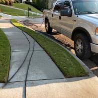 A silver truck parked on the street with a garden hose extending across the concrete sidewalk toward a residential lawn.