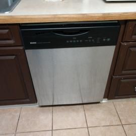 A stainless steel dishwasher with a black control panel is installed between dark wood cabinets in a kitchen.