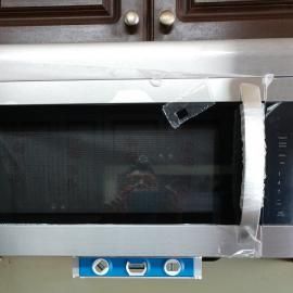 A stainless steel microwave with protective plastic film and a blue level tool attached to the bottom.