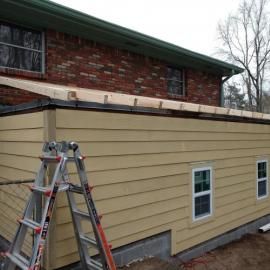 A construction site showing a tan home addition being built against a brick house with a ladder in the foreground.