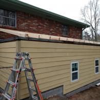 A ladder stands against a tan, siding-covered wall under a brick house with a newly installed wooden roof frame.