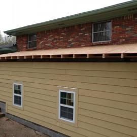 A beige-sided home addition with a newly installed plywood roof under a red brick house wall.